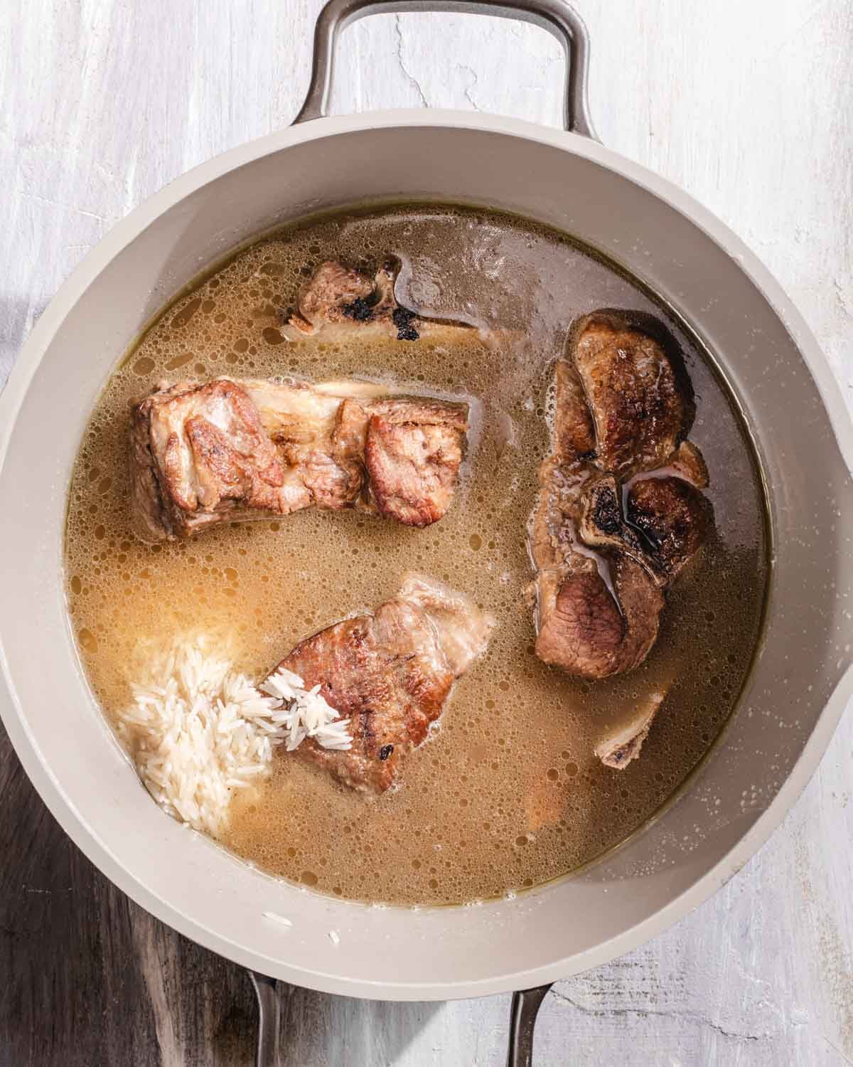 A pot filled with broth, several browned meat pieces, and a small amount of uncooked white rice on the side, all set on a light-colored surface.