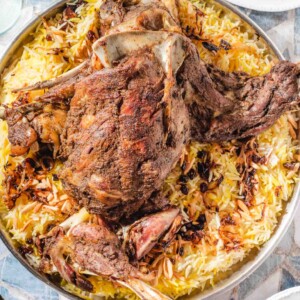 A large serving platter holds roasted lamb atop yellow and white rice, garnished with slivered almonds. Small bowls of green sauce and extra plates are visible around the platter.
