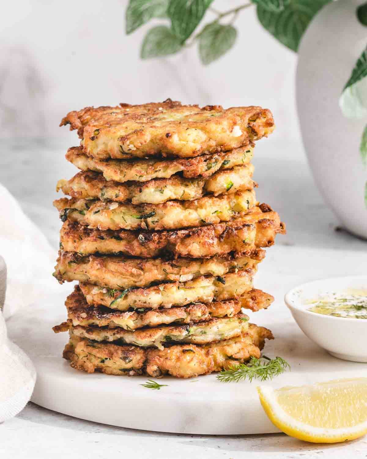 A stack of golden, crispy fritters sits on a round white board next to a bowl of dip, fresh herbs, and a lemon wedge, with a leafy plant in the background.