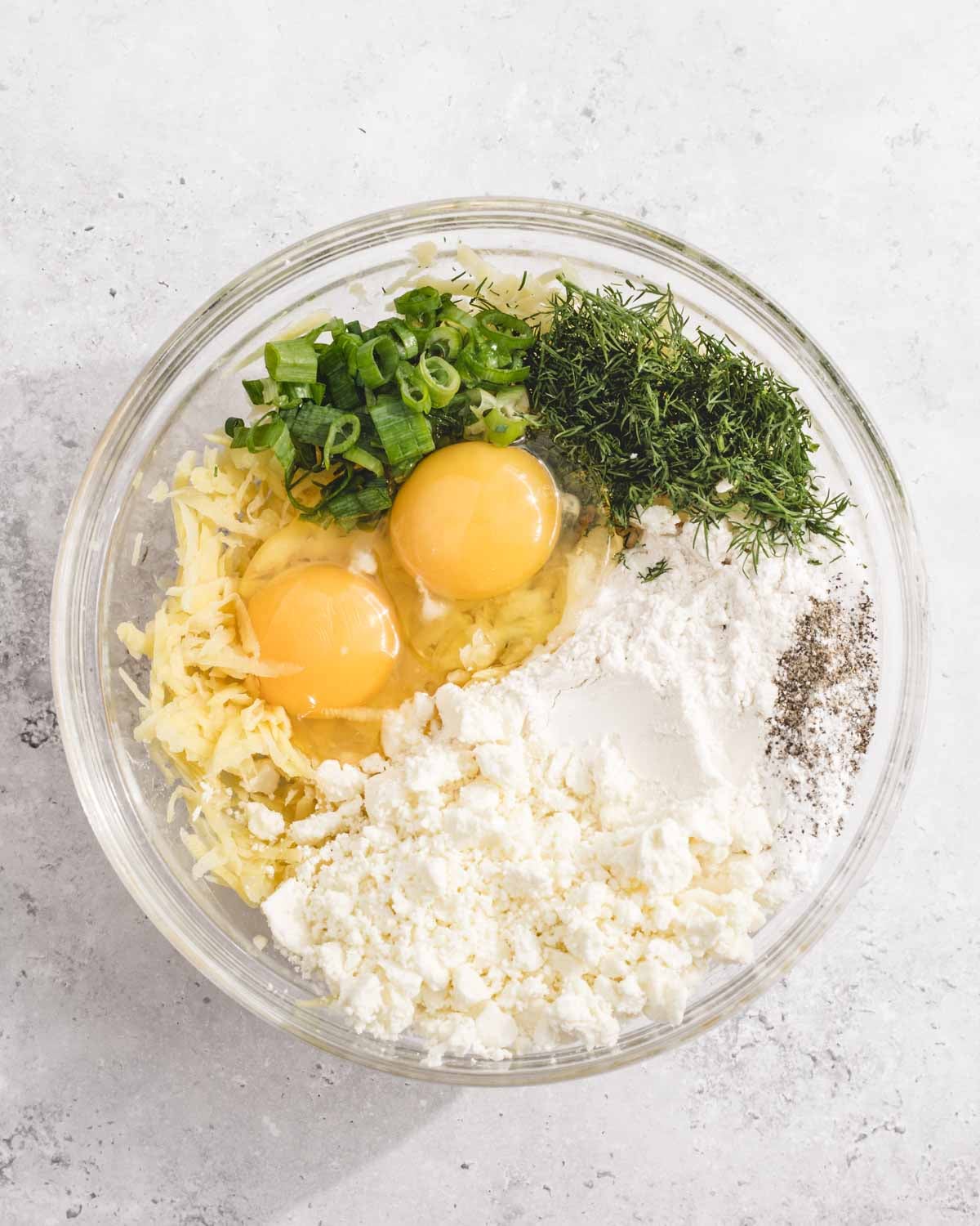 A glass bowl containing eggs, shredded cheese, chopped green onions, fresh dill, flour, feta cheese, and ground black pepper, all arranged separately before mixing.