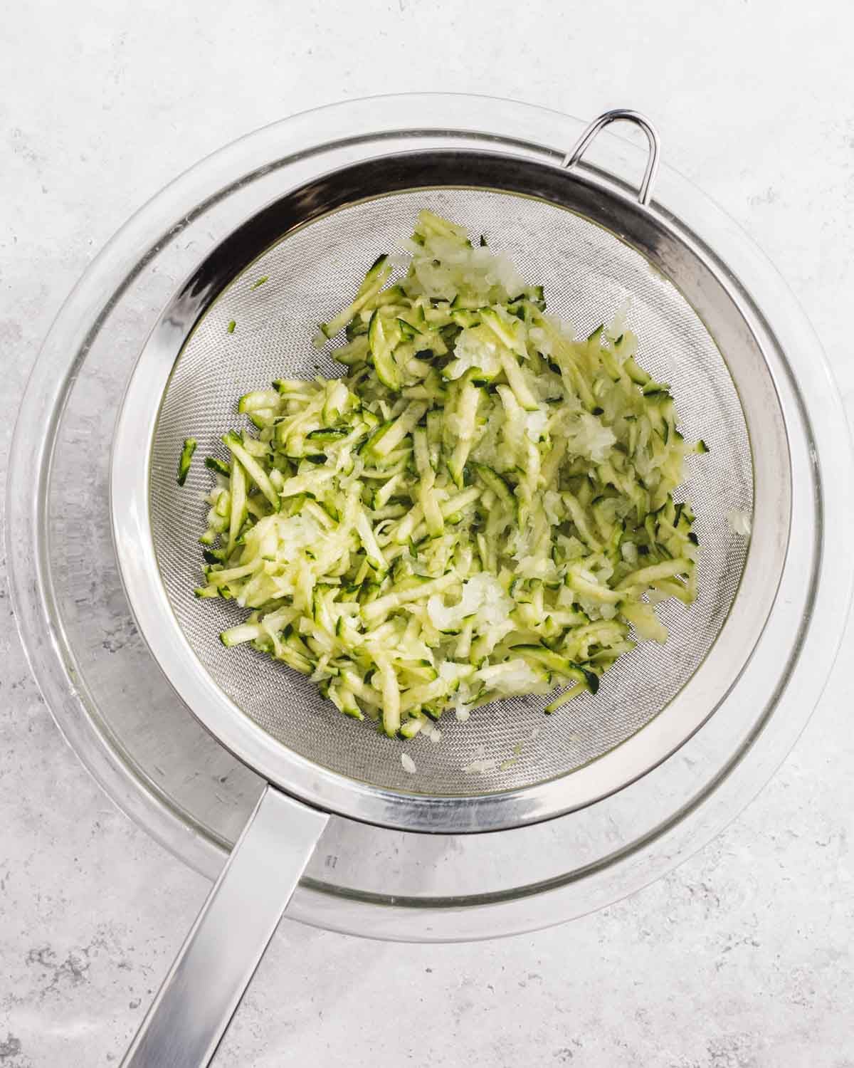 A metal strainer with shredded zucchini rests over a glass bowl on a white surface. The zucchini appears to be draining or drying.
