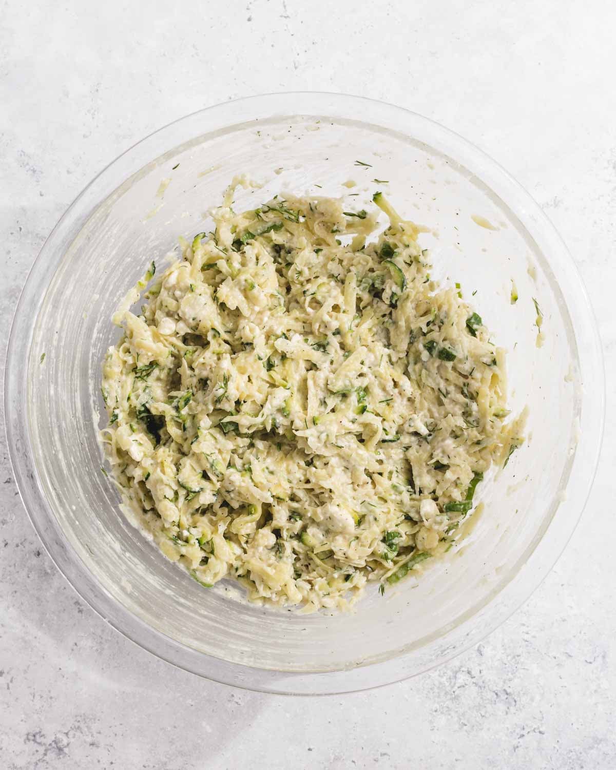 A clear glass bowl filled with a mixture of shredded zucchini, herbs, and cheese sits on a light, textured surface. The mixture appears moist and well combined, ready for cooking or baking.