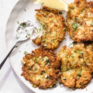 A plate with four golden-brown fritters garnished with chopped herbs, a lemon wedge, and a spoonful of creamy dipping sauce on the side.