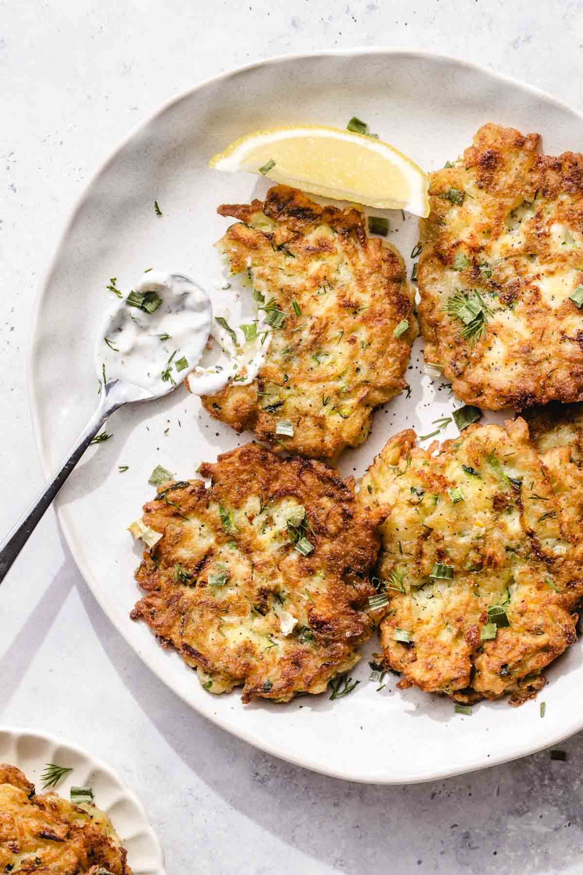 A plate with four golden-brown fritters garnished with chopped herbs, a lemon wedge, and a spoonful of creamy dipping sauce on the side.