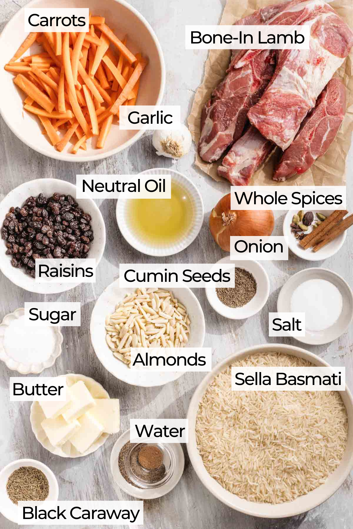 Various labeled ingredients for a recipe are arranged on a light surface, including bone-in lamb, carrots, garlic, basmati rice, whole spices, onion, oil, raisins, cumin seeds, almonds, salt, sugar, butter, water, and black caraway.