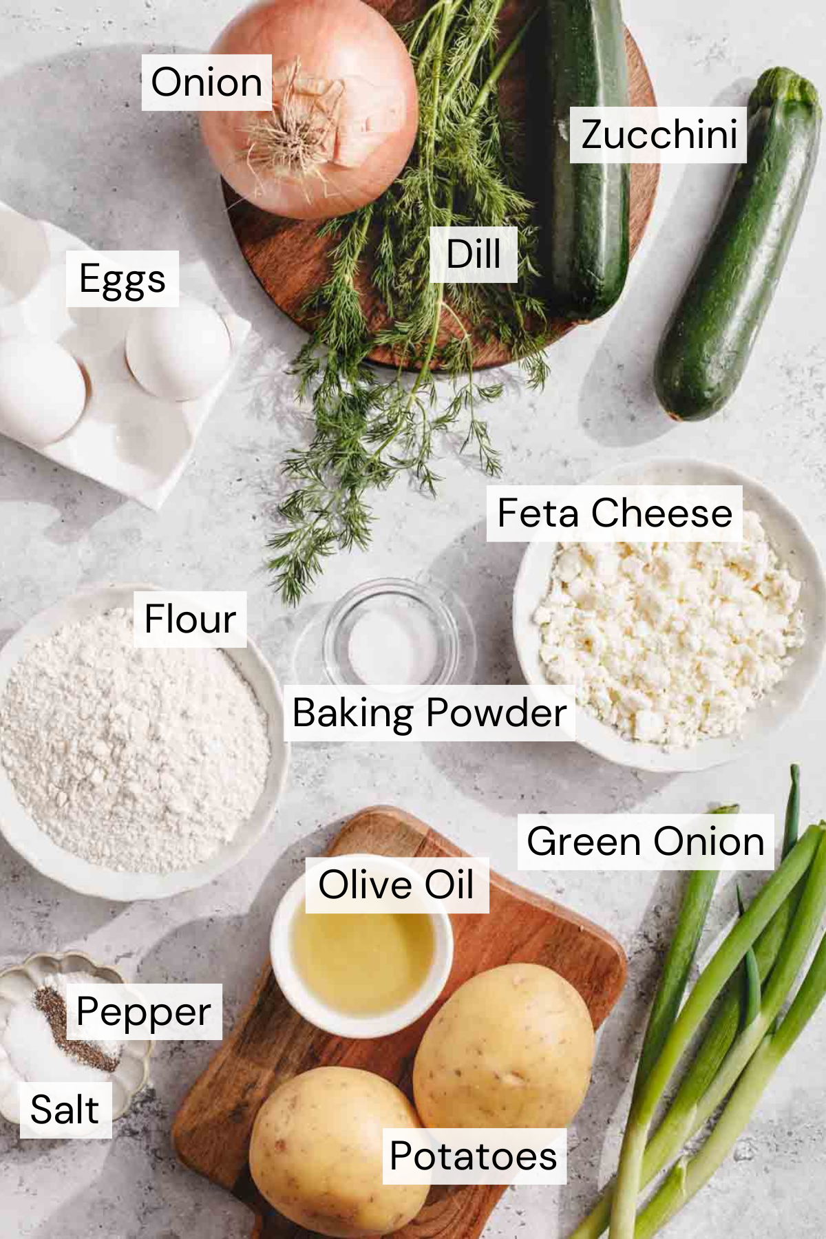 Top-down view of various labeled ingredients on a light surface, including onion, zucchini, eggs, dill, flour, feta cheese, baking powder, green onion, olive oil, potatoes, salt, and pepper.
