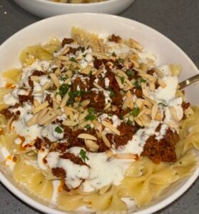 A bowl of Turkish pasta bowtie topped with ground meat sauce, a white creamy sauce, slivered almonds, and chopped parsley on a gray countertop.