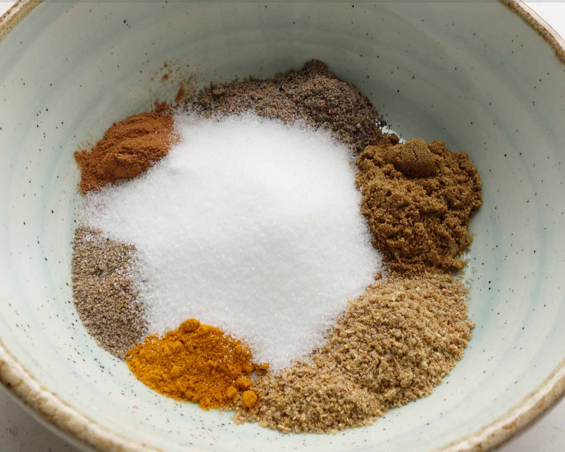 A light blue bowl containing separate piles of ground spices, including white granulated sugar, turmeric, black pepper, cinnamon, and other brown and tan spices.