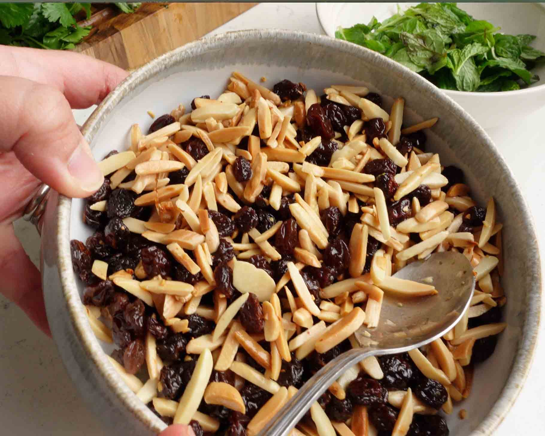 A hand holds a bowl filled with slivered almonds and raisins, with a metal spoon inside. In the background, there is a white bowl containing fresh green herbs or lettuce leaves.