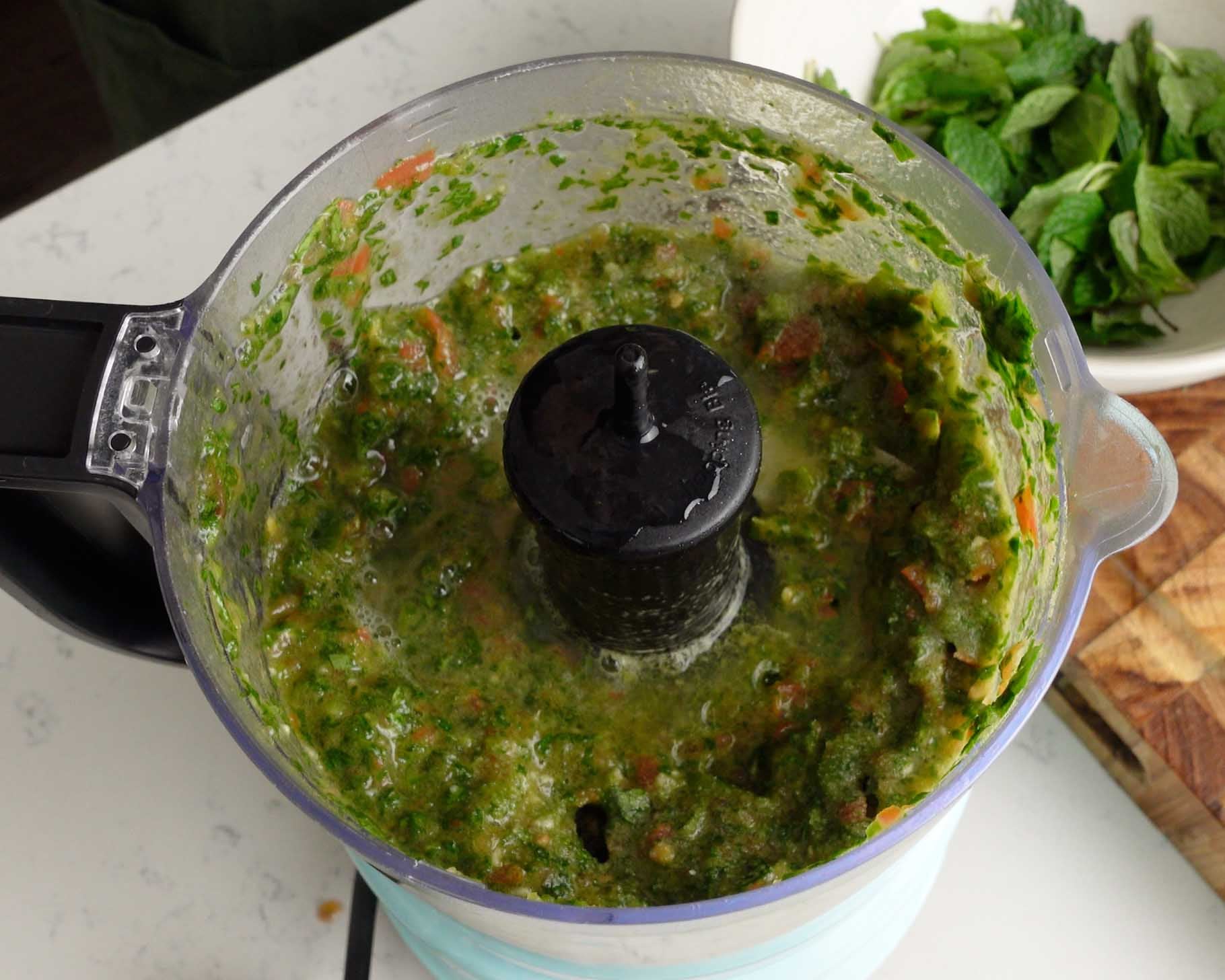 A food processor containing a green, chunky mixture sits on a counter. Fresh mint leaves and a wooden cutting board are visible in the background.
