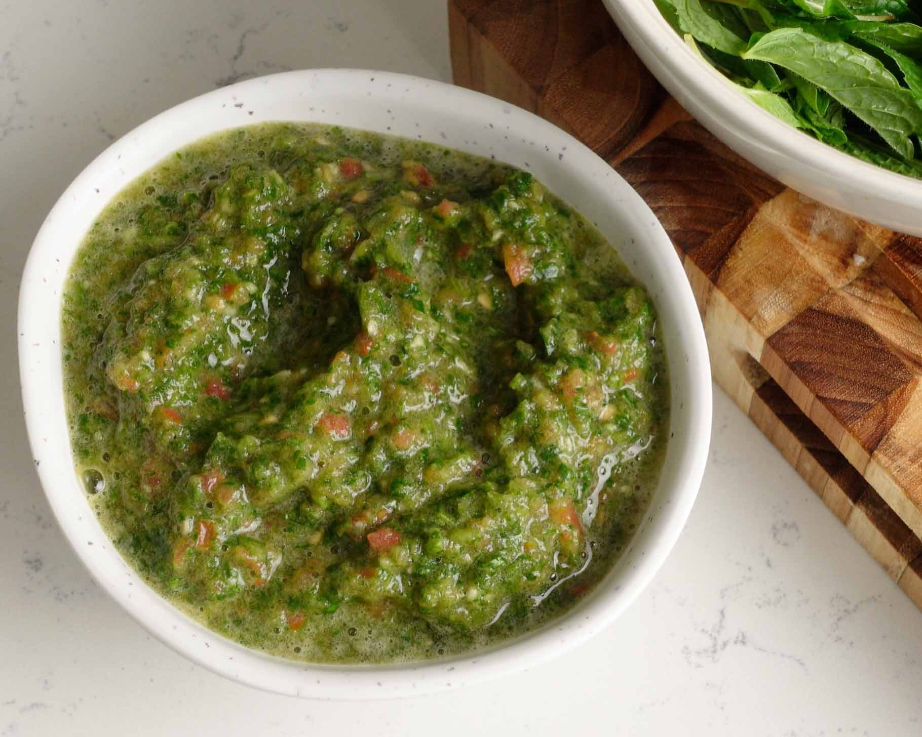 A white bowl filled with chunky green sauce, placed on a light countertop next to a wooden cutting board and a bowl of leafy greens.