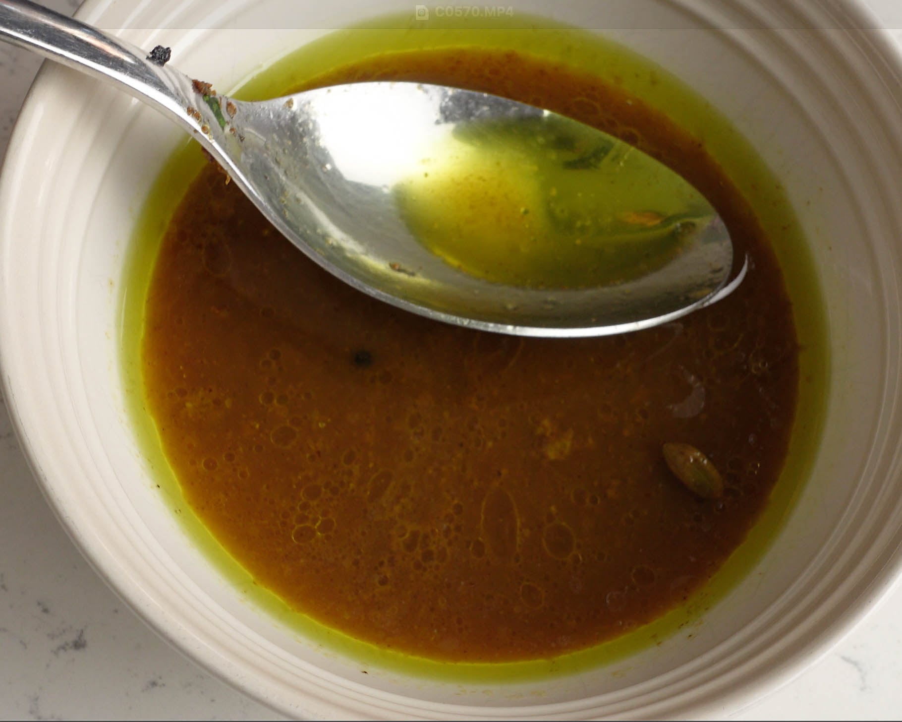 A close-up of a white bowl containing an oily, golden-brown liquid with visible spices. A metal spoon rests inside, reflecting the green-tinted oil on its surface.