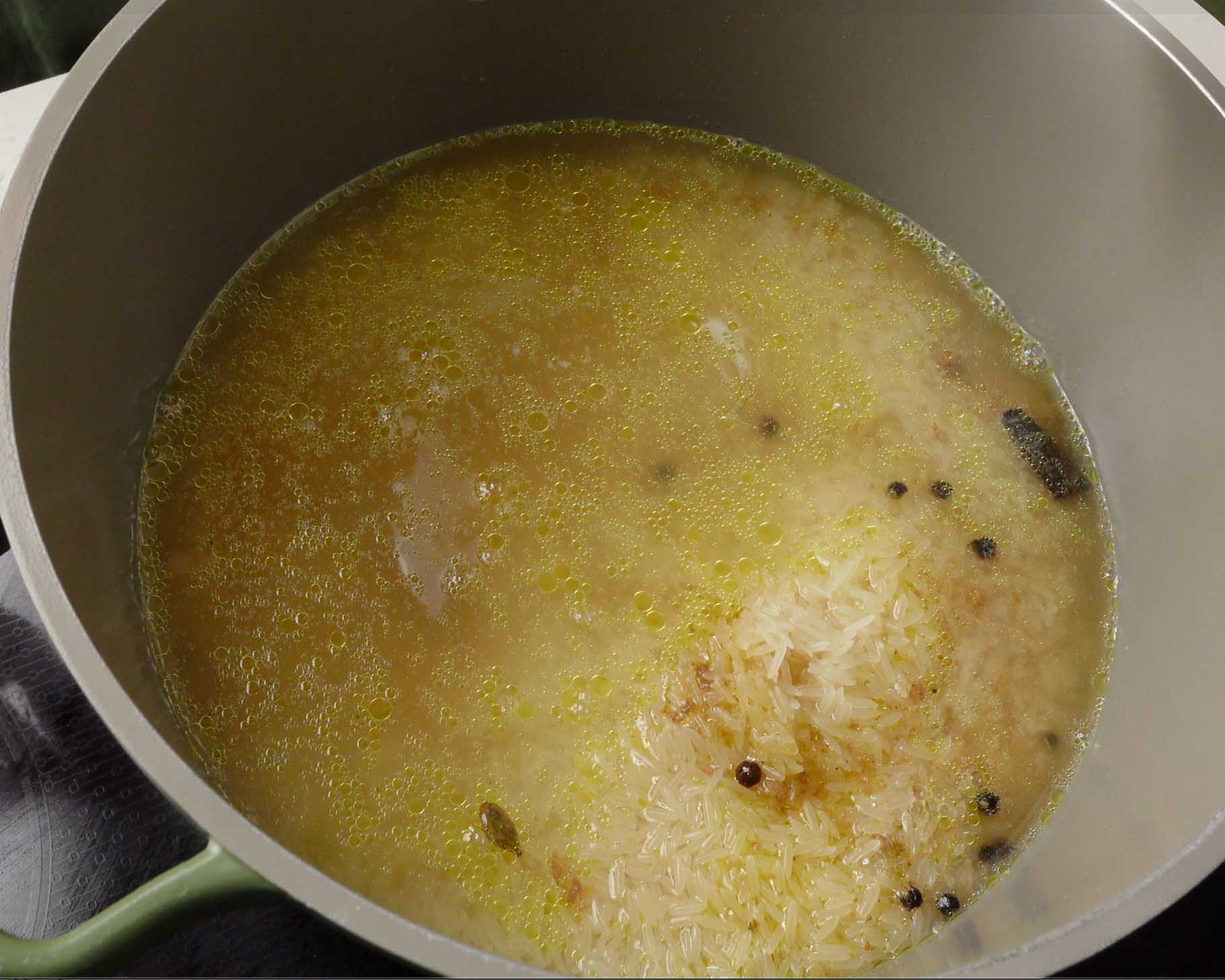 A pot filled with uncooked rice, whole spices, oil, and water, sitting on a stove and starting to cook. Some rice is clumped together, and oil droplets are visible on the surface.