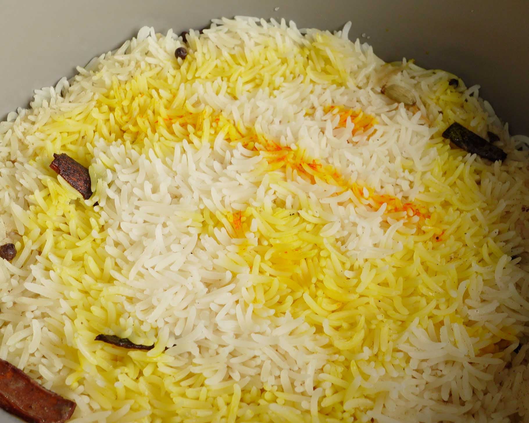 A close-up of cooked basmati rice with a mix of white and yellow grains, garnished with whole spices like cinnamon sticks and cloves.