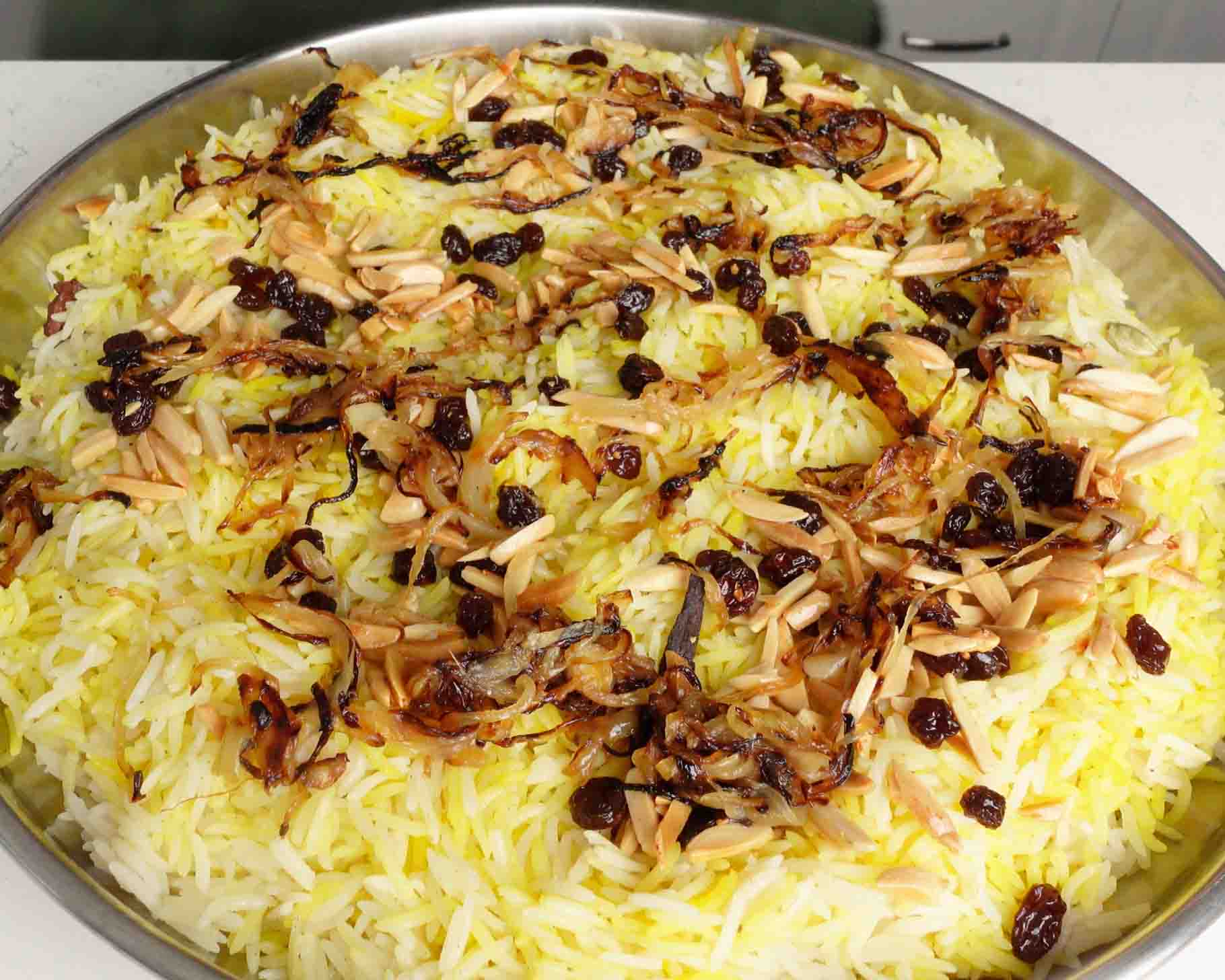 A large round tray of saffron rice garnished with caramelized onions, raisins, and slivered almonds. The rice is fluffy and yellow, with toppings scattered evenly across the surface.