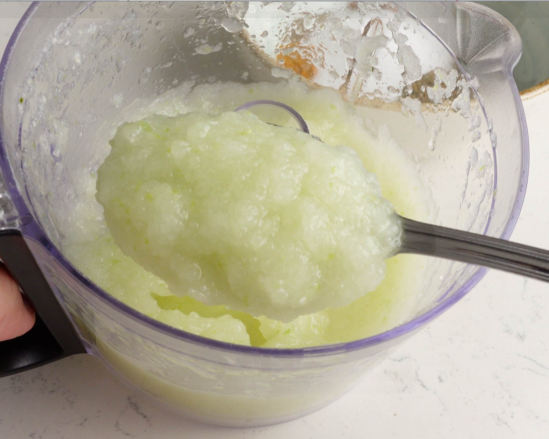 A spoon scooping a thick, pale green, slushy mixture from a transparent blender jug on a white countertop.