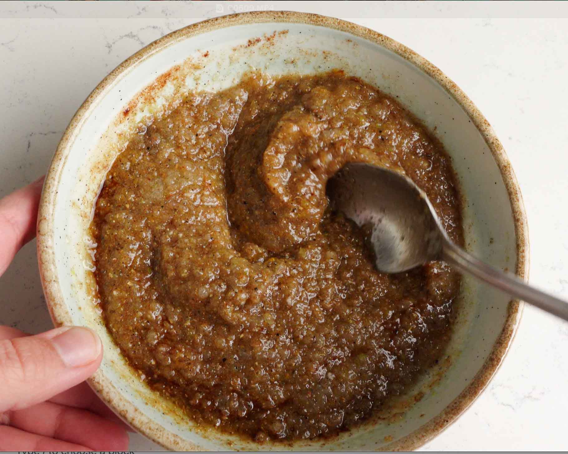 A hand holds a bowl filled with a thick, brown, grainy mixture while a spoon scoops some of it. The bowl sits on a light-colored surface.