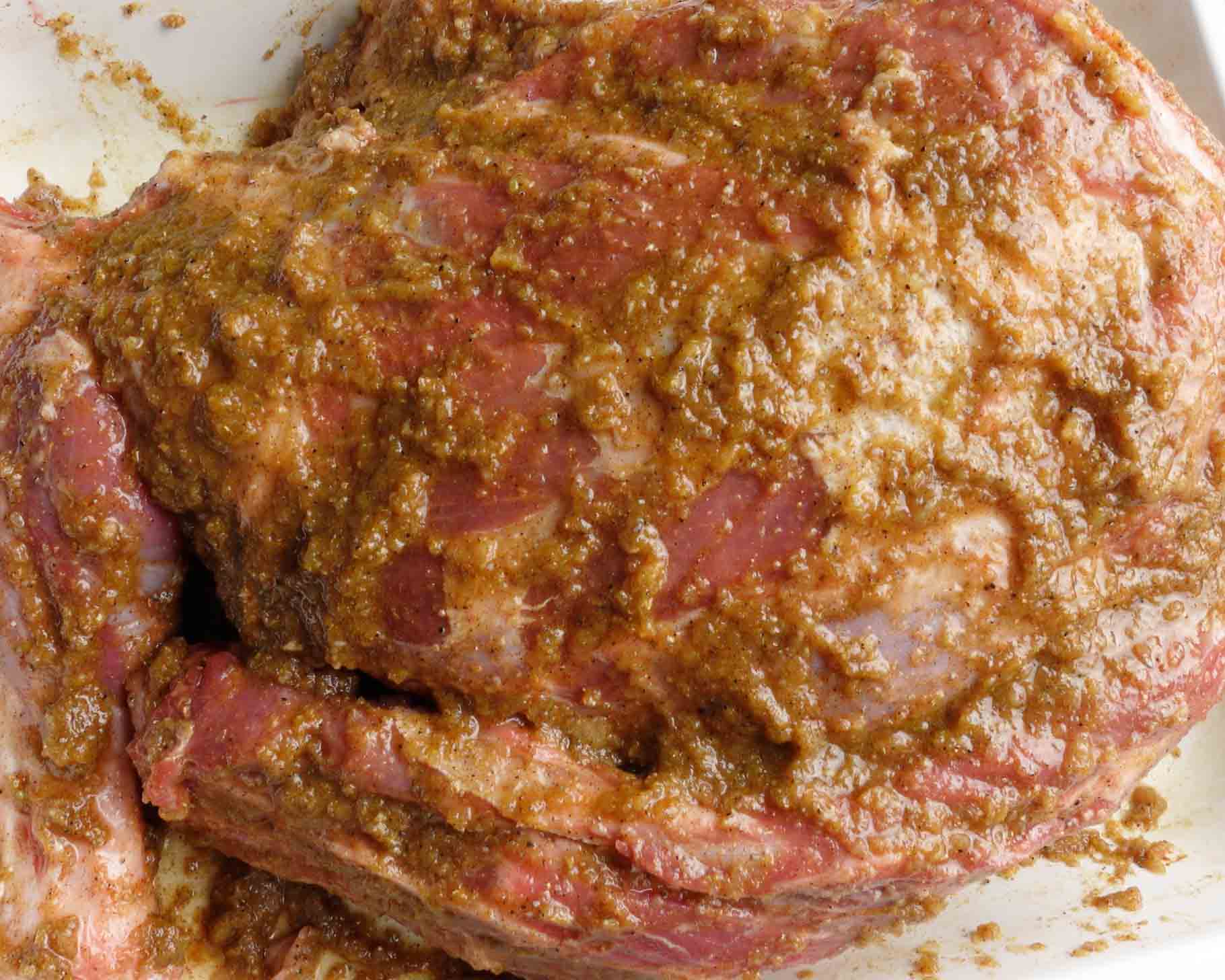 Close-up of a large piece of raw lamb coated with a thick, brownish marinade or spice rub, resting in a white dish.