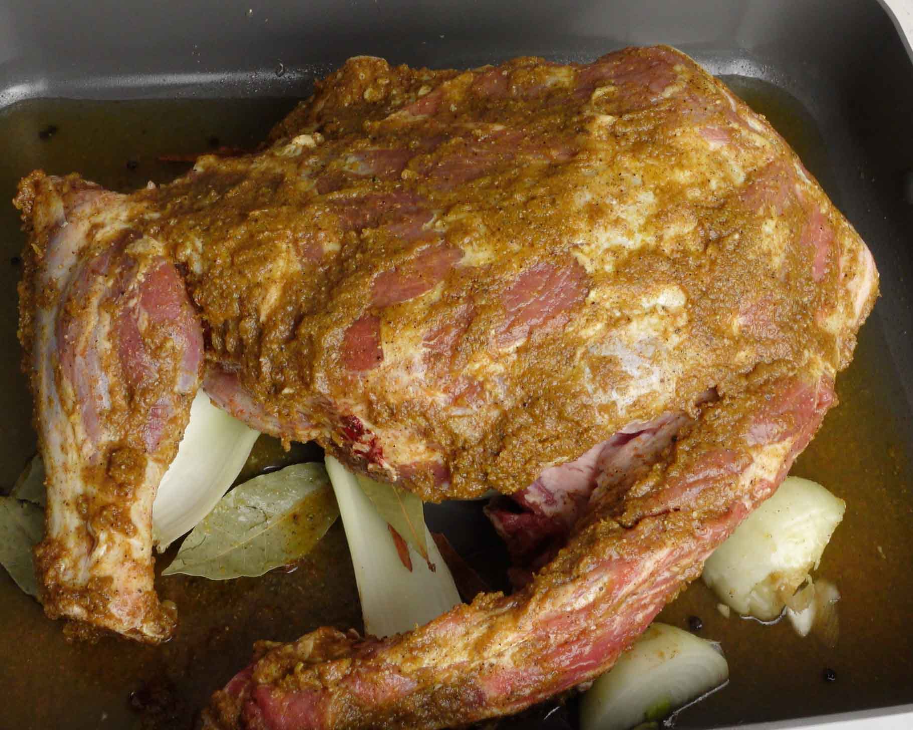 A marinated piece of lamb, rests on chopped onions and bay leaves in a roasting pan, ready to be cooked.