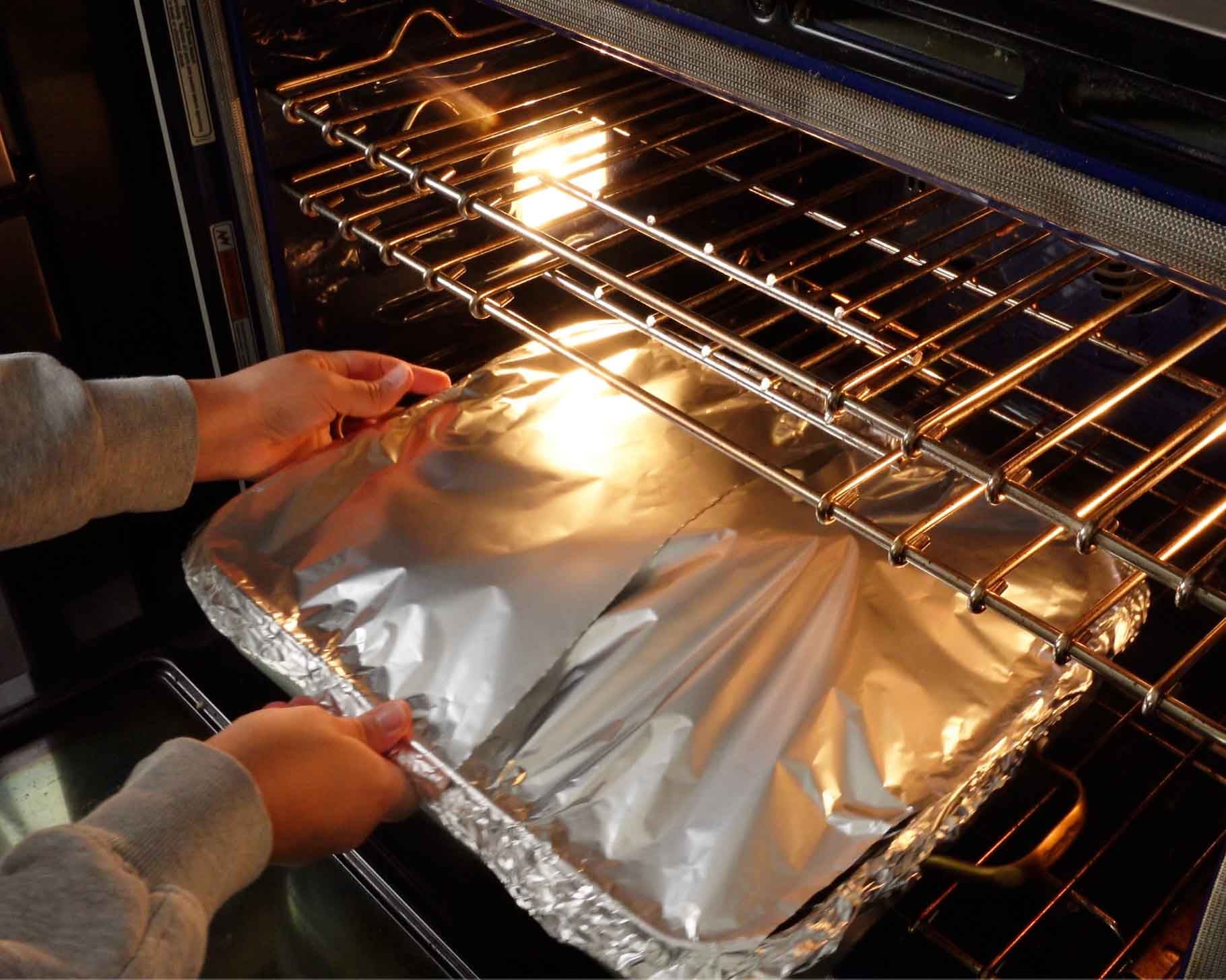A person is placing a large, foil-covered baking dish onto the middle rack of an oven, with the oven light on and both hands holding the dish.