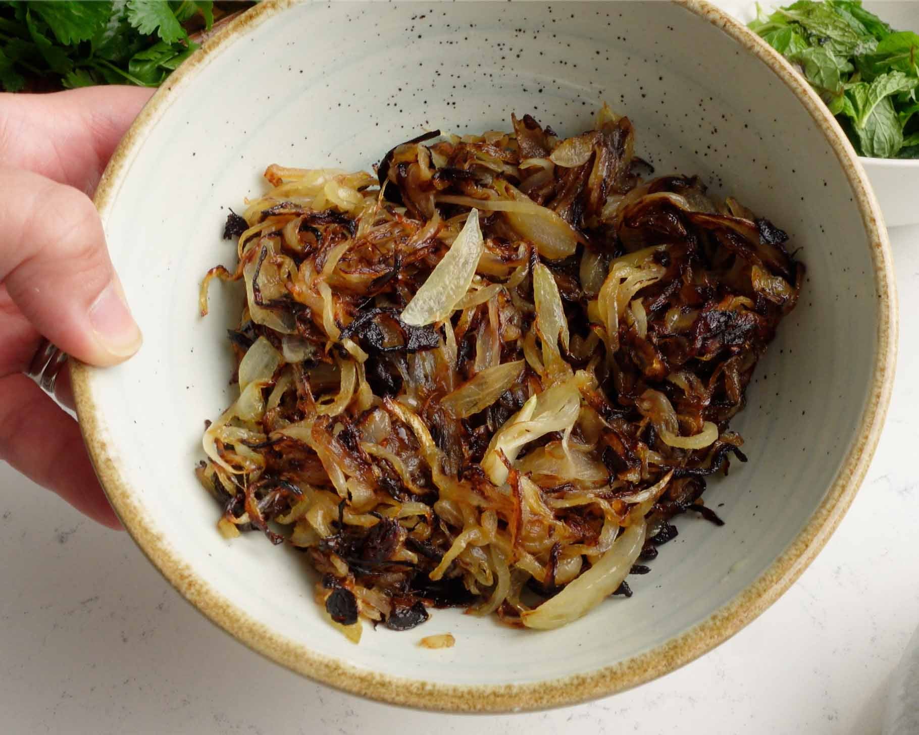 A hand holds a speckled ceramic bowl filled with caramelized onions, golden brown and slightly charred, on a light countertop with greens visible in the background.