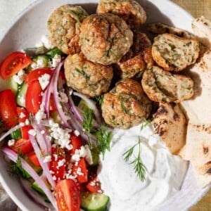 A bowl with Greek meatballs, sliced pita bread, tzatziki sauce, and a salad made of cherry tomatoes, cucumber, red onion, feta cheese, and fresh dill, all arranged on a white plate over a yellow and white tablecloth.