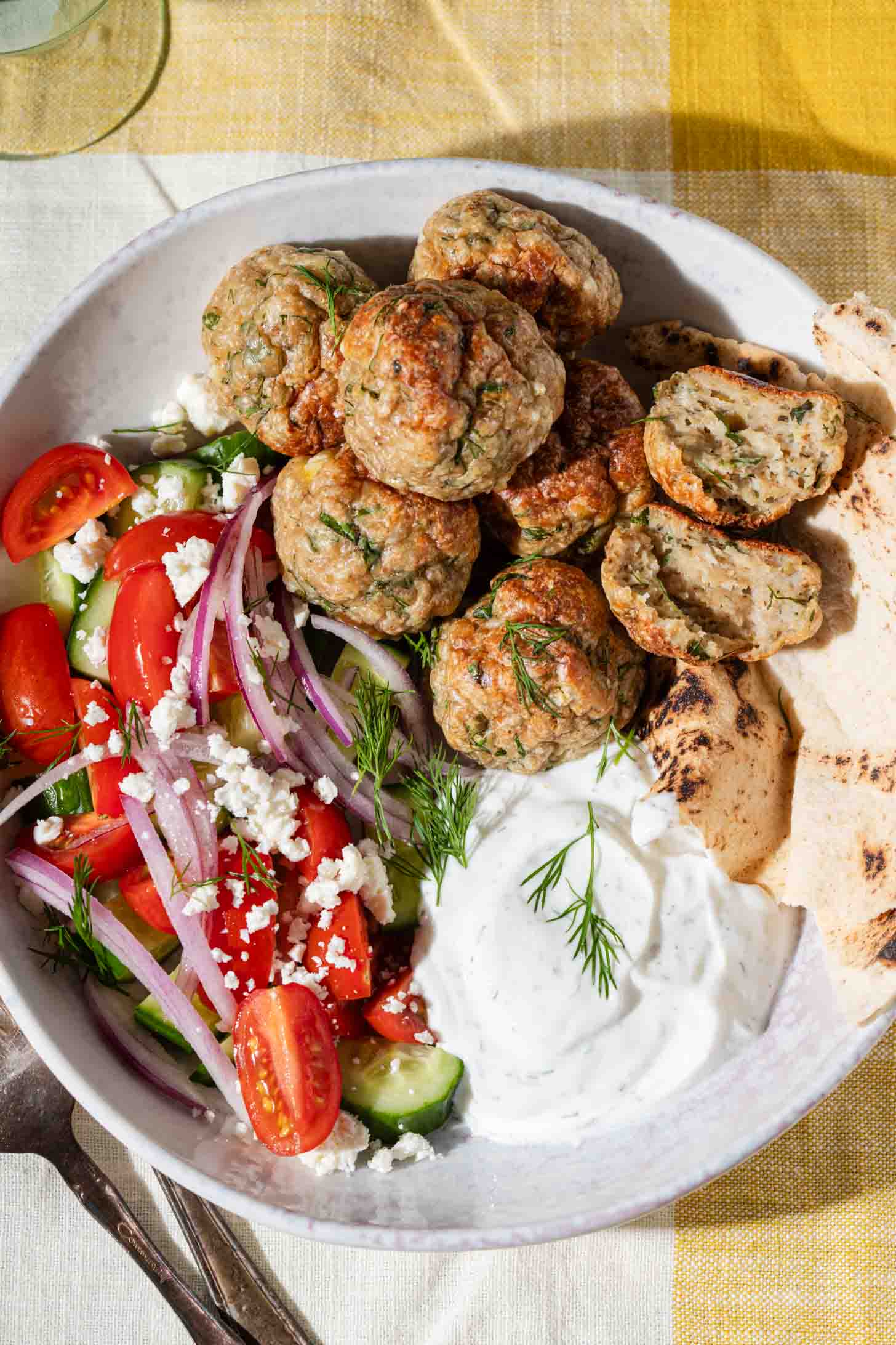 A bowl with Greek meatballs, sliced pita bread, tzatziki sauce, and a salad made of cherry tomatoes, cucumber, red onion, feta cheese, and fresh dill, all arranged on a white plate over a yellow and white tablecloth.