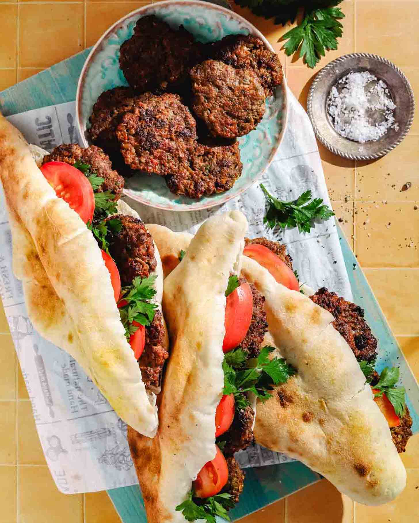 Three sandwiches filled with grilled meat patties, tomato slices, and parsley on flatbread sit on a board next to a plate of extra meat patties and a small bowl of coarse salt.