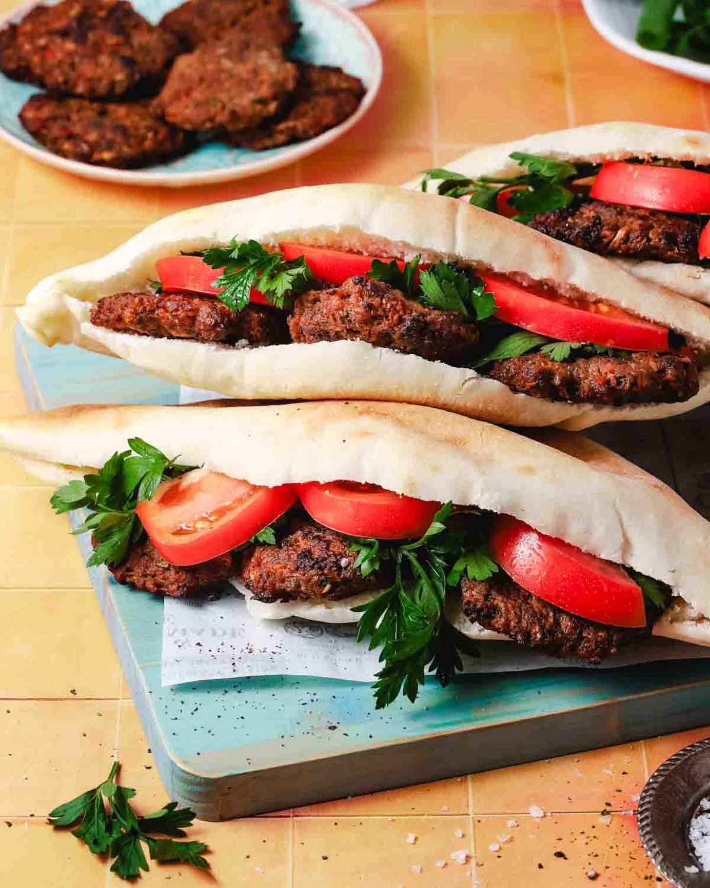 Two pita sandwiches filled with falafel patties, fresh tomato slices, and parsley are placed on a wooden board. More falafel and greens are visible in the background.