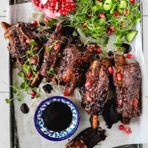 A tray with glazed ribs garnished with microgreens and pomegranate seeds, next to cucumber pieces, a halved pomegranate, a bowl of dark sauce, and a sauce brush on parchment paper.
