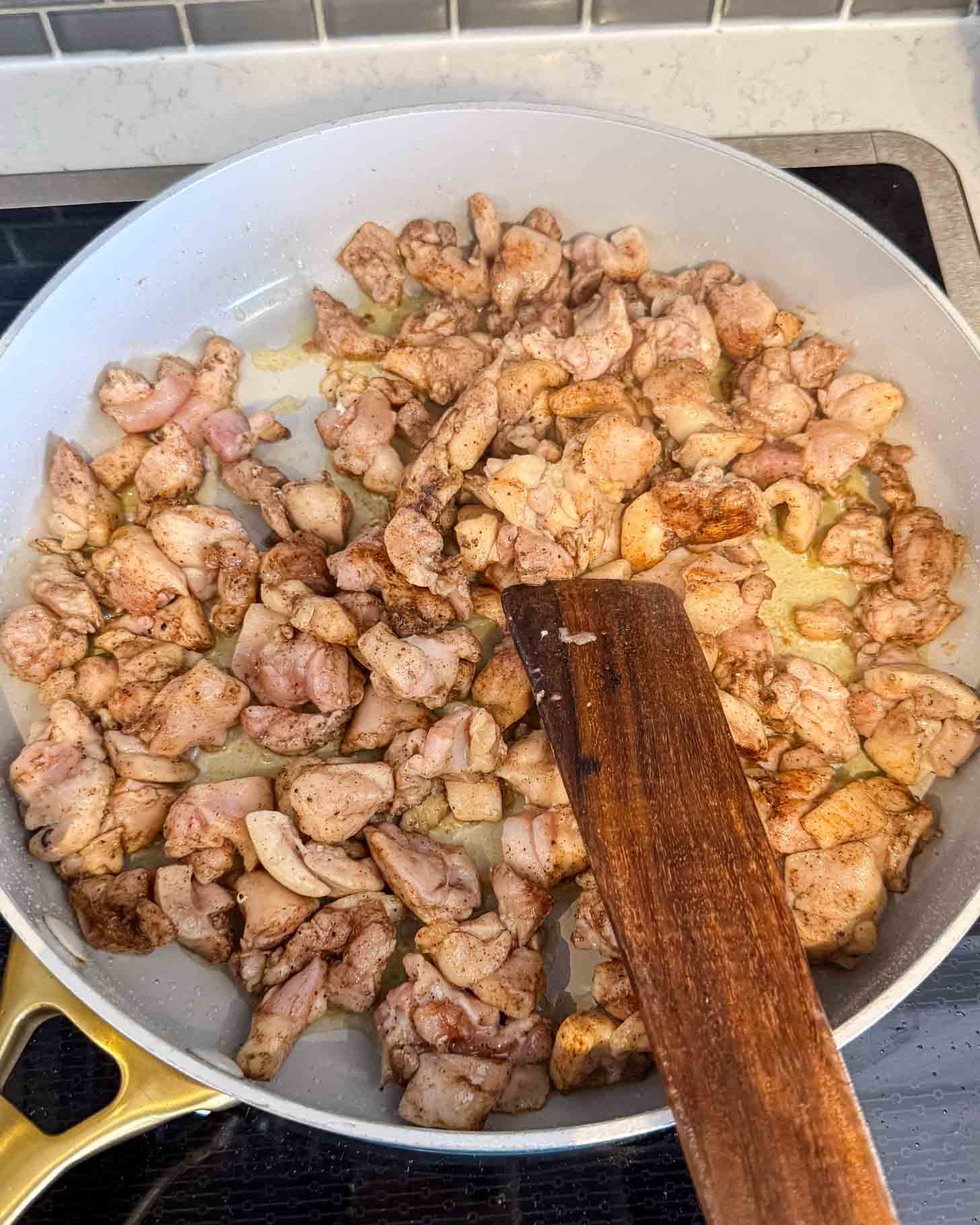 A frying pan filled with seasoned, diced pieces of raw chicken being cooked on a stovetop. A wooden spatula is resting in the pan, stirring the chicken. White subway tile backsplash is visible in the background.