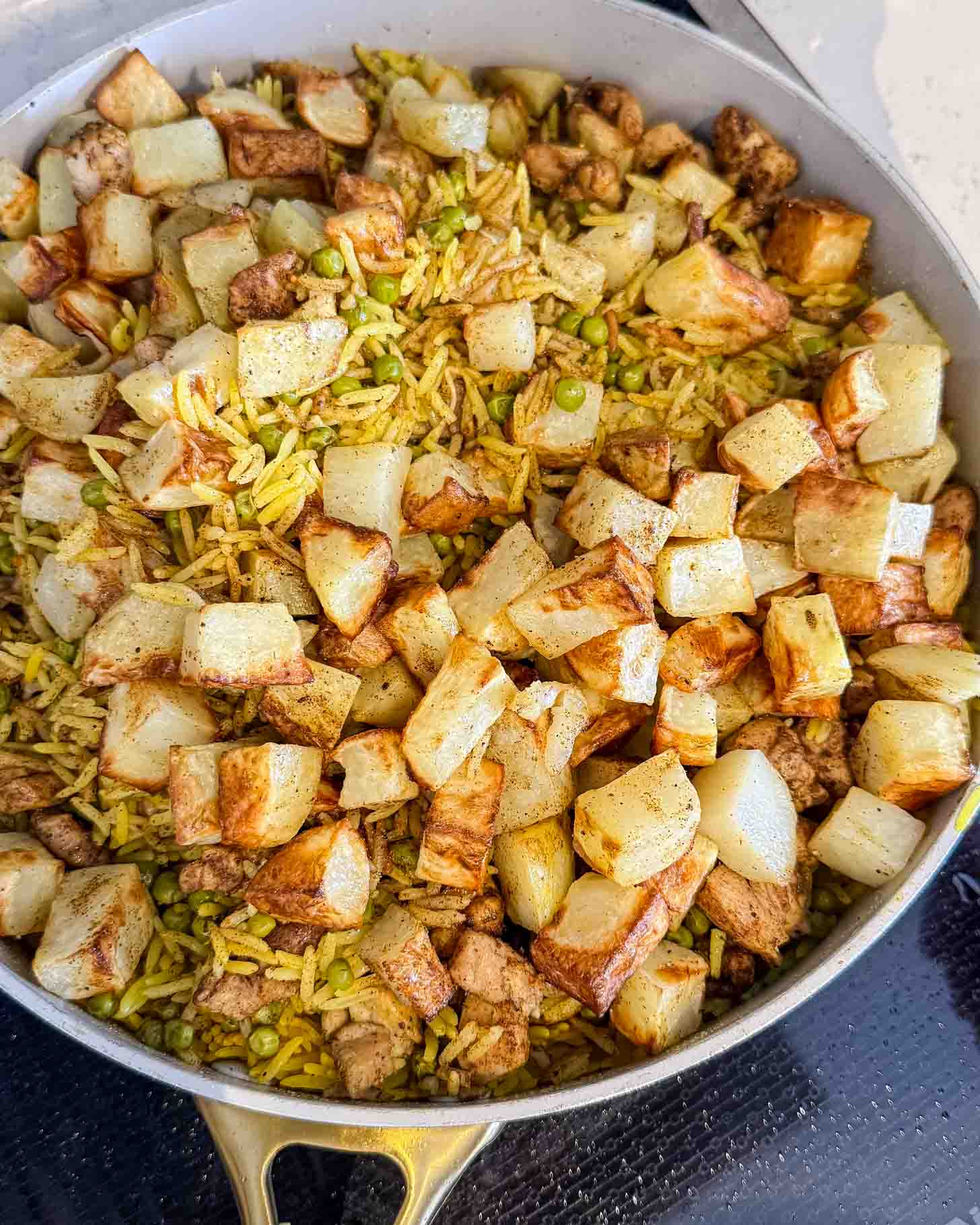 A pan filled with yellow rice, green peas, chunks of cooked chicken, and golden-brown roasted potato cubes. The ingredients are mixed together, creating a colorful and hearty dish.