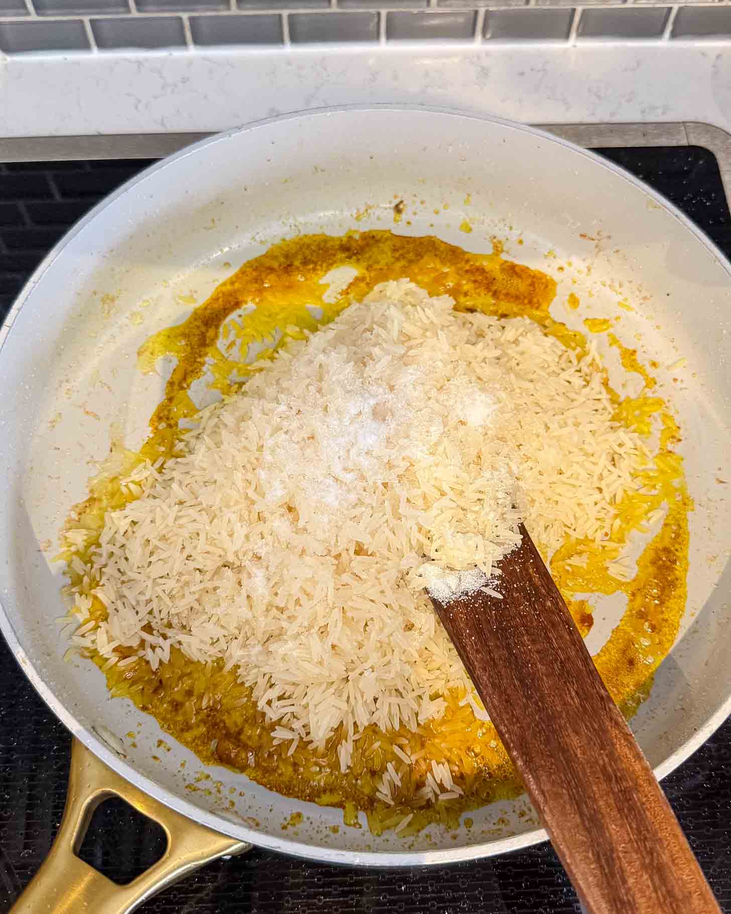 Uncooked white rice and seasoning are being stirred in a white skillet with a wooden spatula on a stovetop. The skillet contains yellow spices and rice is being combined with them.