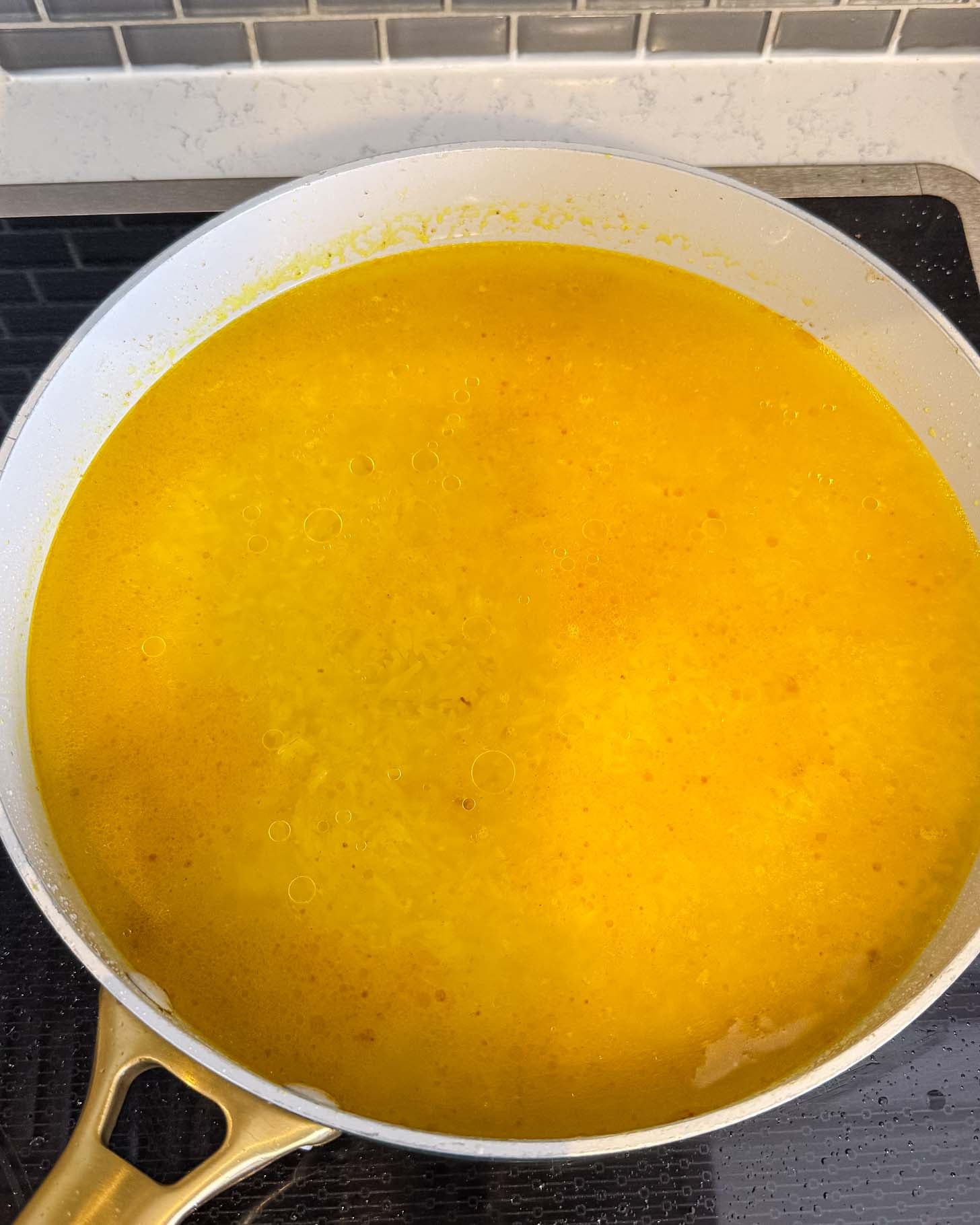 A large white pan on a stovetop is filled with yellow liquid and rice, simmering and bubbling as it cooks. The bright color suggests the presence of turmeric or saffron.