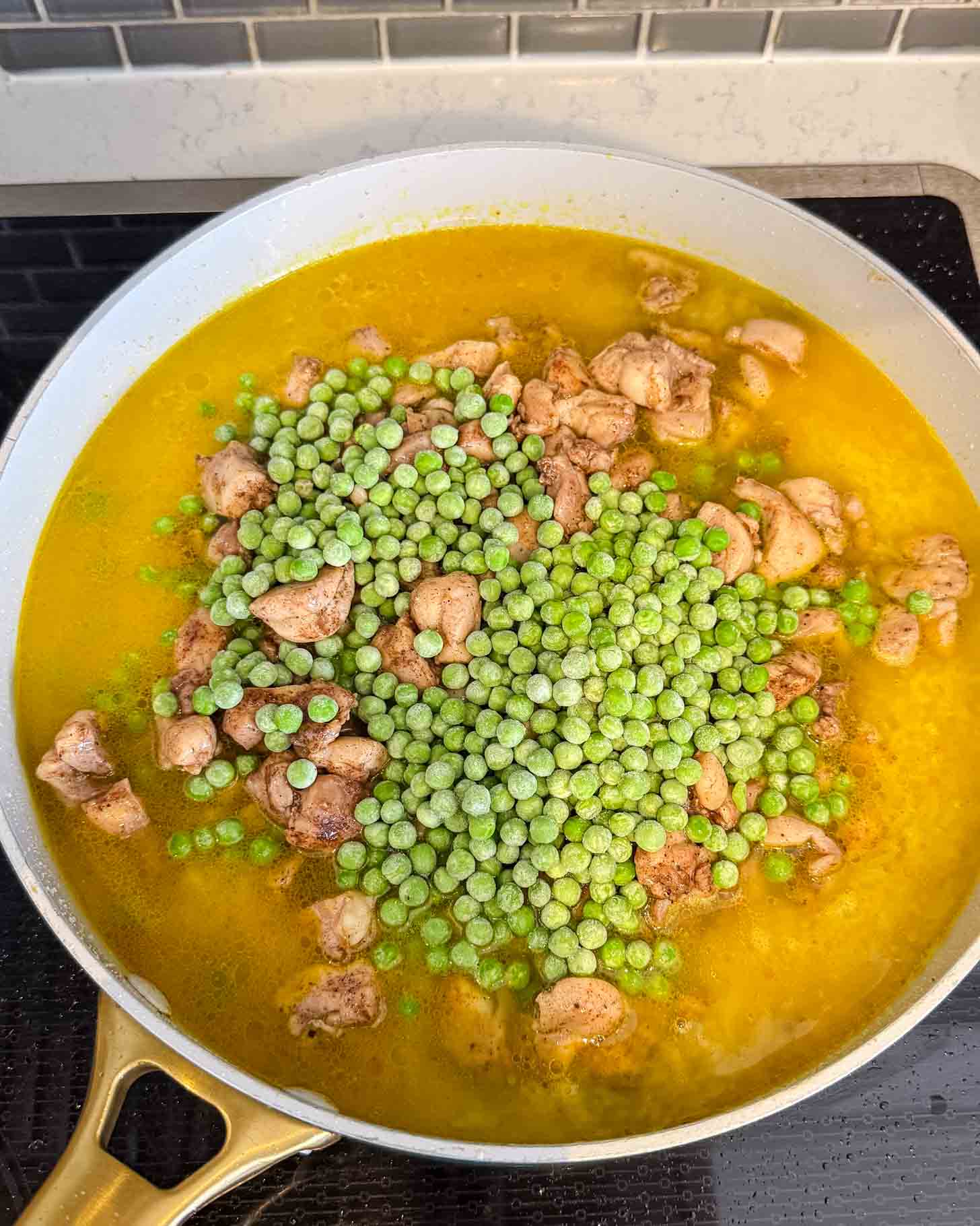 A skillet on a stovetop contains simmering chicken pieces in a yellow broth, topped with a heap of frozen green peas.