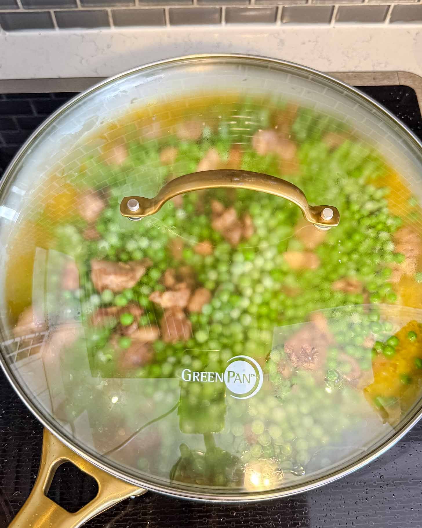 A glass-lidded GreenPan skillet on a stovetop, showing steamed-up condensation with visible green peas and chunks of brown meat cooking inside.