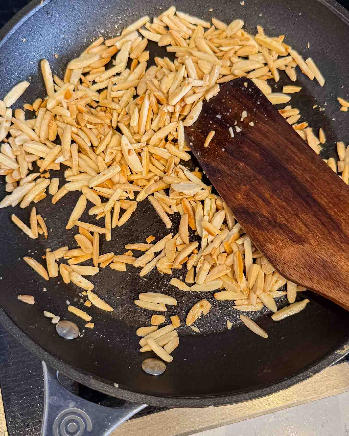 Slivered almonds being toasted in a black frying pan with a wooden spatula, some pieces lightly browned.