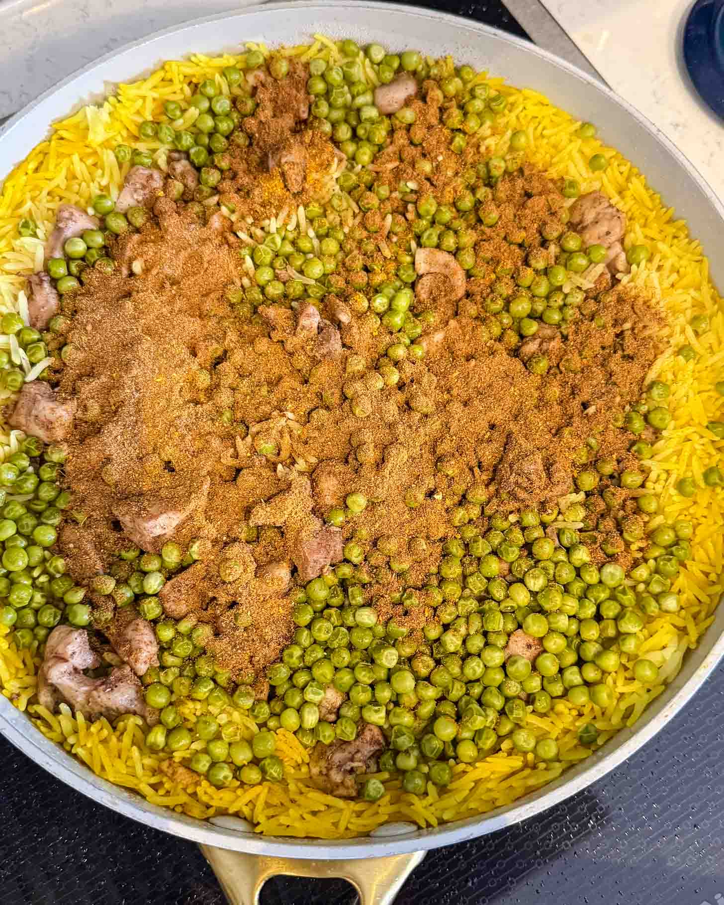 A skillet filled with yellow rice, green peas, pieces of meat, and a generous layer of brown spices spread across the top, ready to be mixed and cooked.