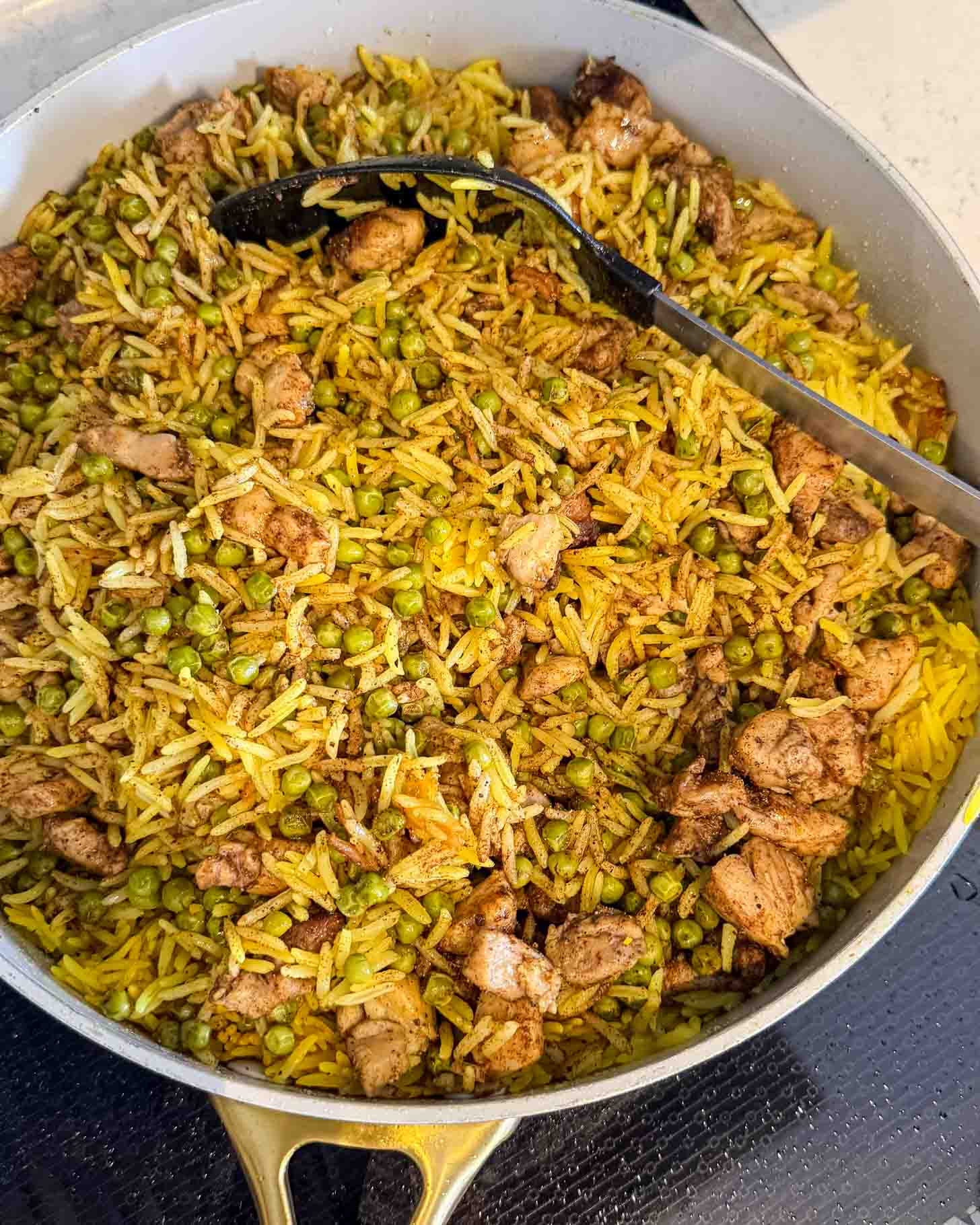 A large pan filled with yellow rice, green peas, and chunks of seasoned chicken, with a black serving spoon resting inside the dish.
