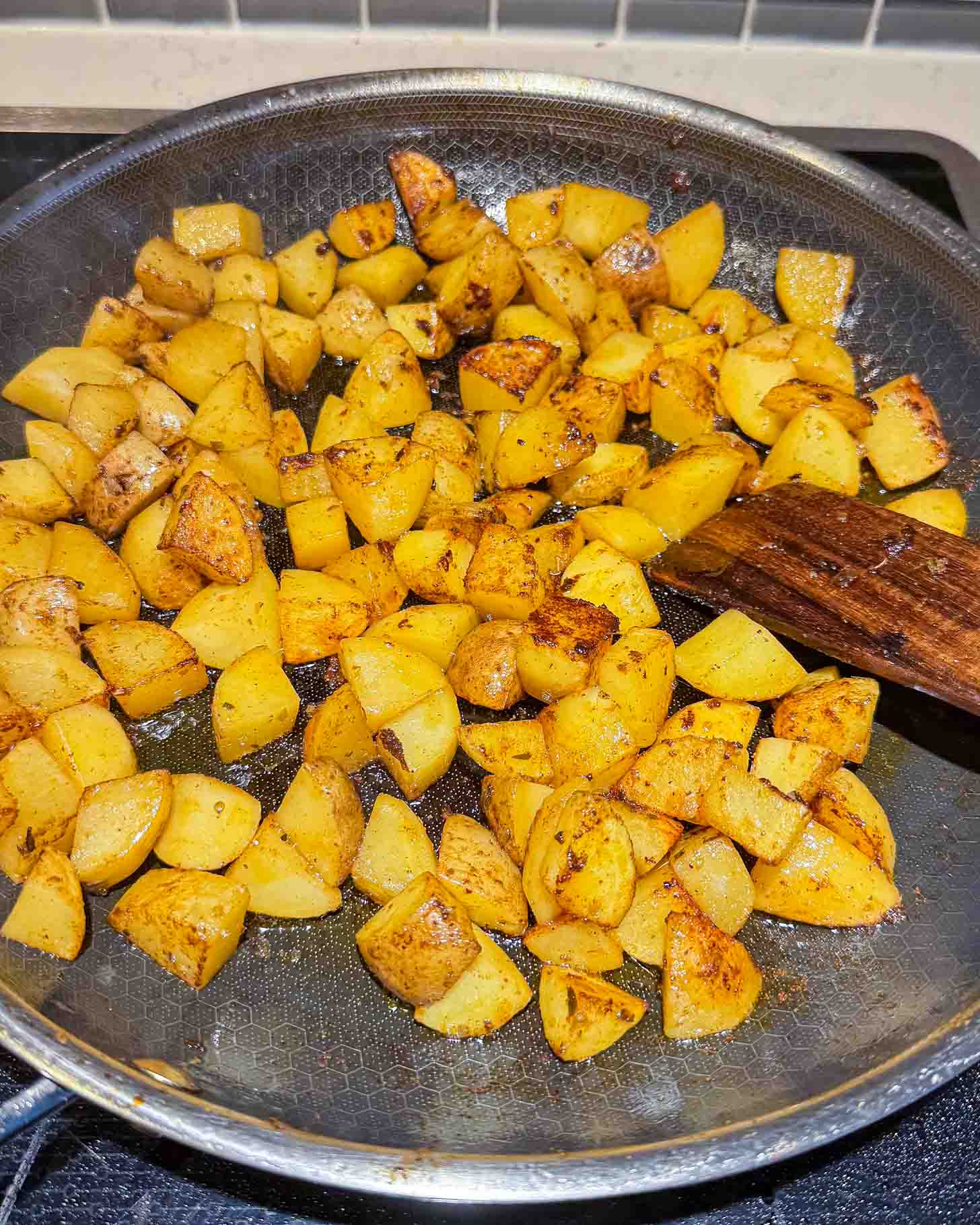 Diced potatoes being sautéed in a frying pan with seasoning; a wooden spatula rests on the side of the pan.