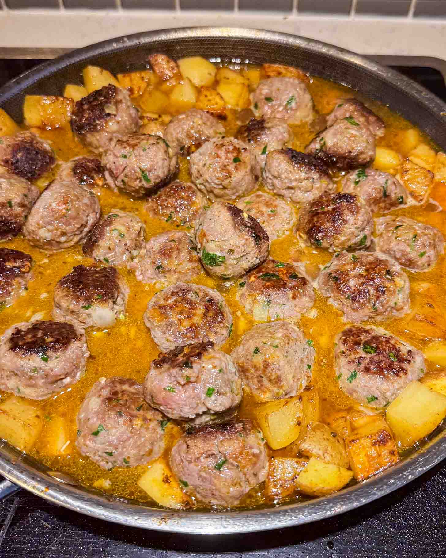 A pan filled with browned meatballs and cubed potatoes simmering in a golden, seasoned sauce on a stovetop.