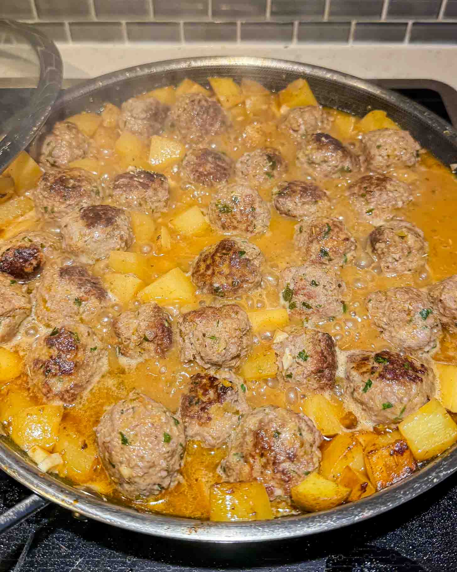 Meatballs and cubed potatoes simmer in a rich brown sauce in a large skillet on a stovetop, with chopped herbs sprinkled on top.