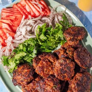 A green plate with grilled meat patties, sliced tomatoes, sliced onions sprinkled with spices, and fresh parsley. Two glasses with light yellow drinks are in the background.