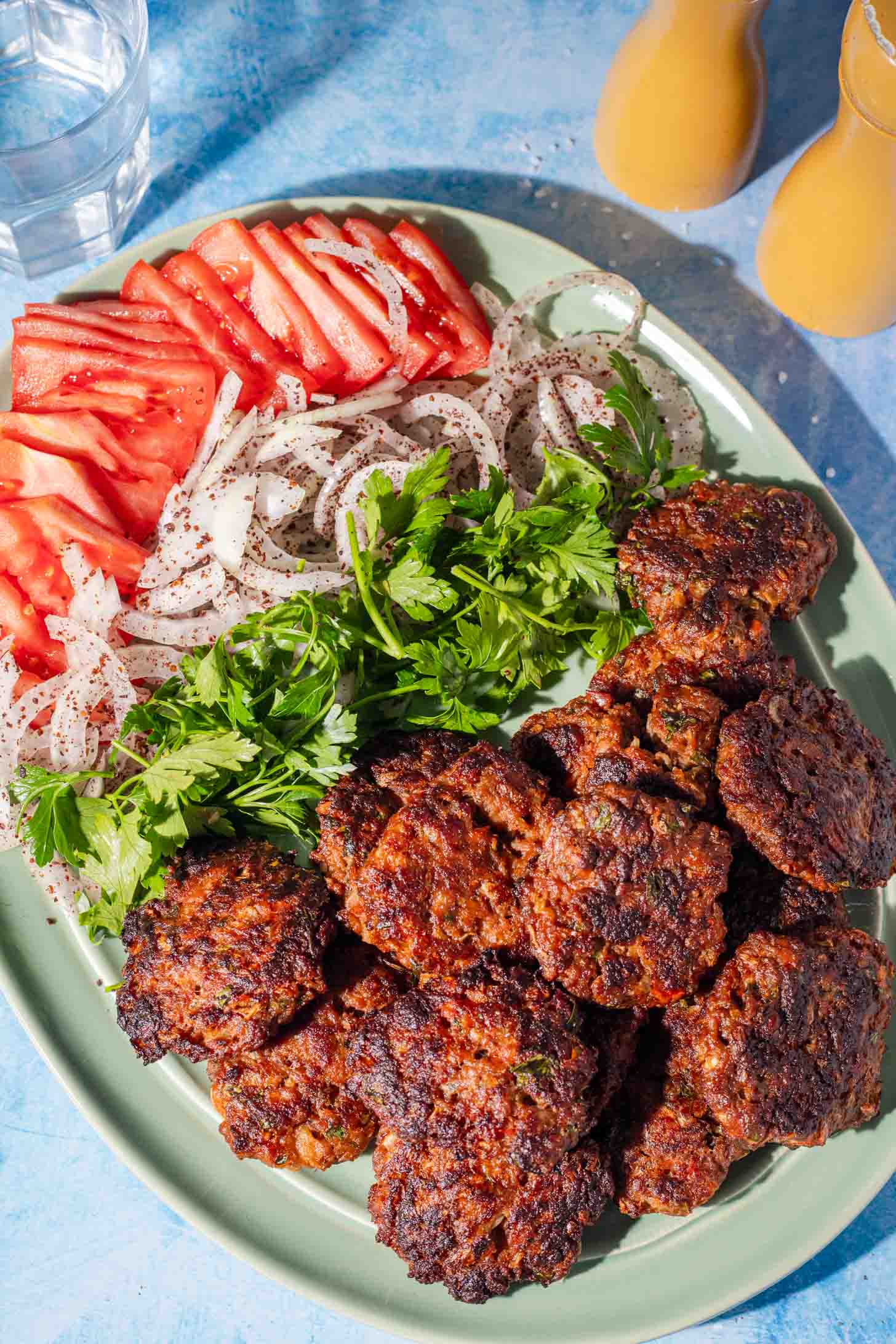 A green plate with grilled meat patties, sliced tomatoes, sliced onions sprinkled with spices, and fresh parsley. Two glasses with light yellow drinks are in the background.