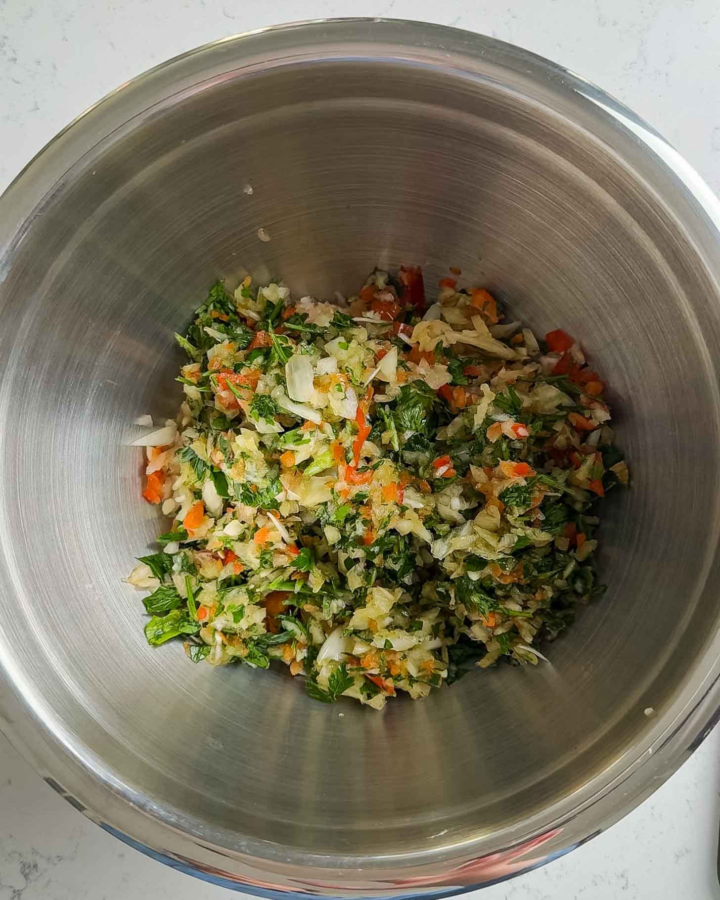 A metal mixing bowl containing a colorful chopped vegetable mixture, including green leafy herbs, orange carrots, red bell peppers, and white cabbage, set on a light-colored countertop.