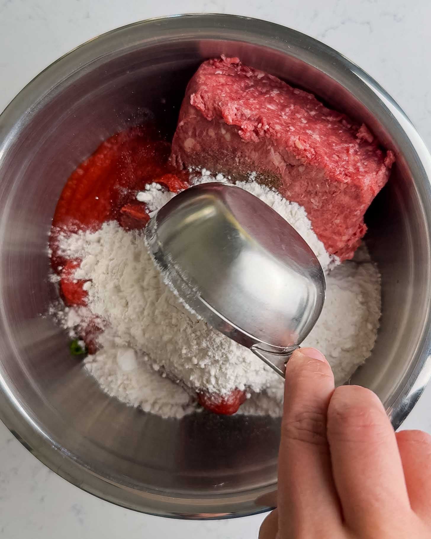 A hand holds a metal measuring cup over a mixing bowl containing ground meat, flour, and a red ingredient, possibly tomato sauce or ketchup, on a white countertop.