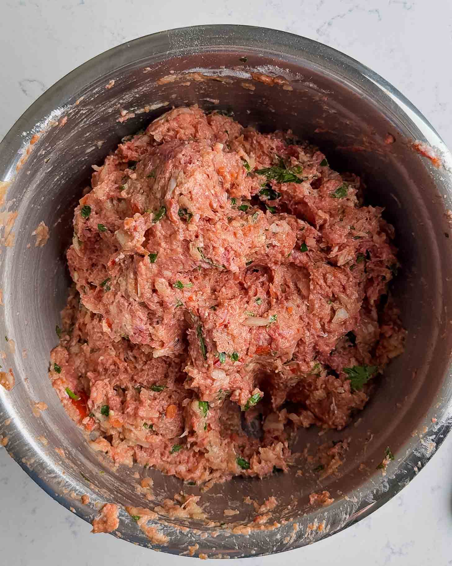 A stainless steel bowl filled with a mixture of raw ground meat, herbs, and seasonings, ready to be prepared for cooking.