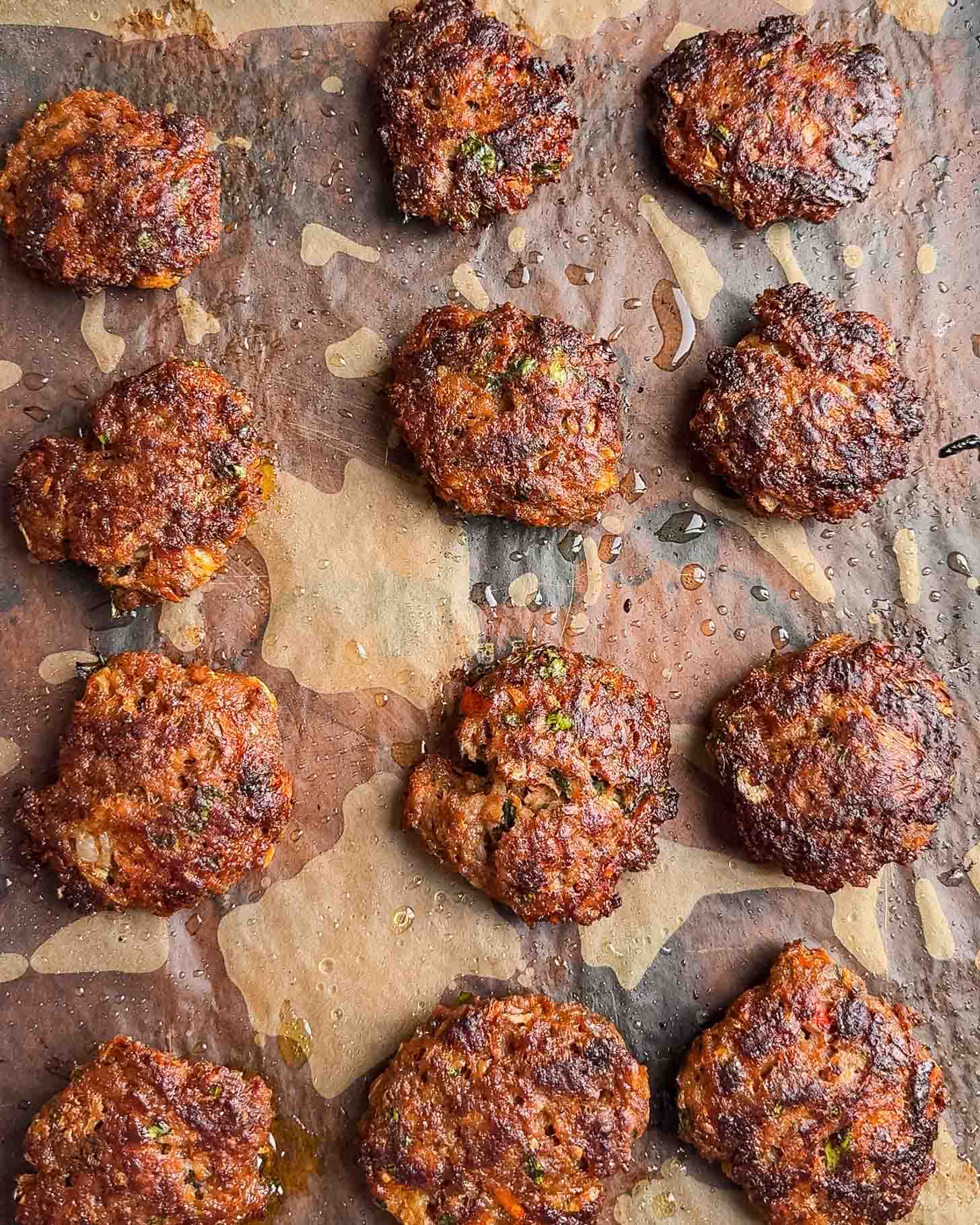 Twelve browned, crispy beef patties are arranged on a baking sheet lined with parchment paper, with visible bits of herbs and small oil spots around them.
