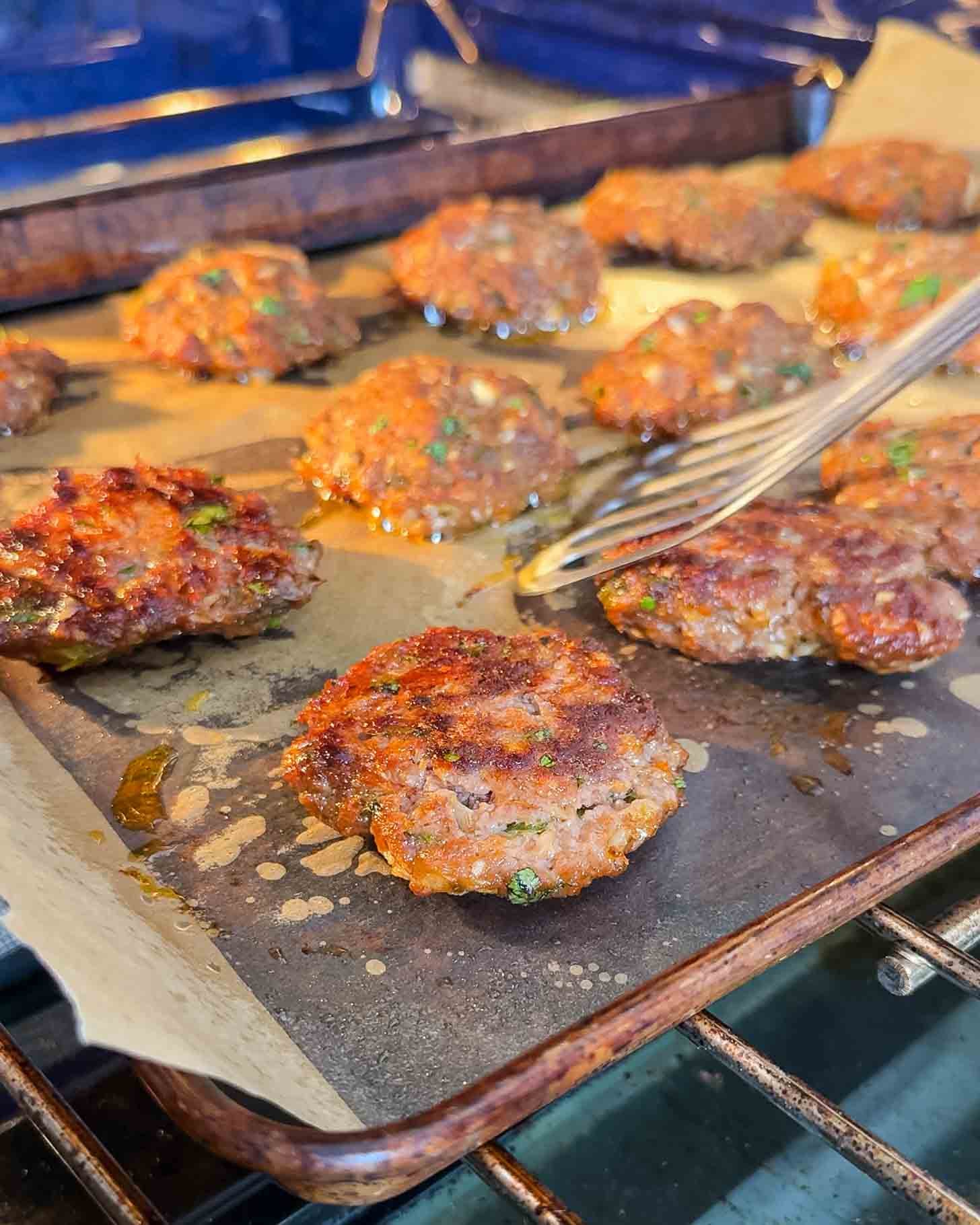 Several browned sausage patties are baking on a parchment-lined baking sheet in an oven. A fork is lifting one of the patties, showing their crispy, cooked texture with some oil around them.