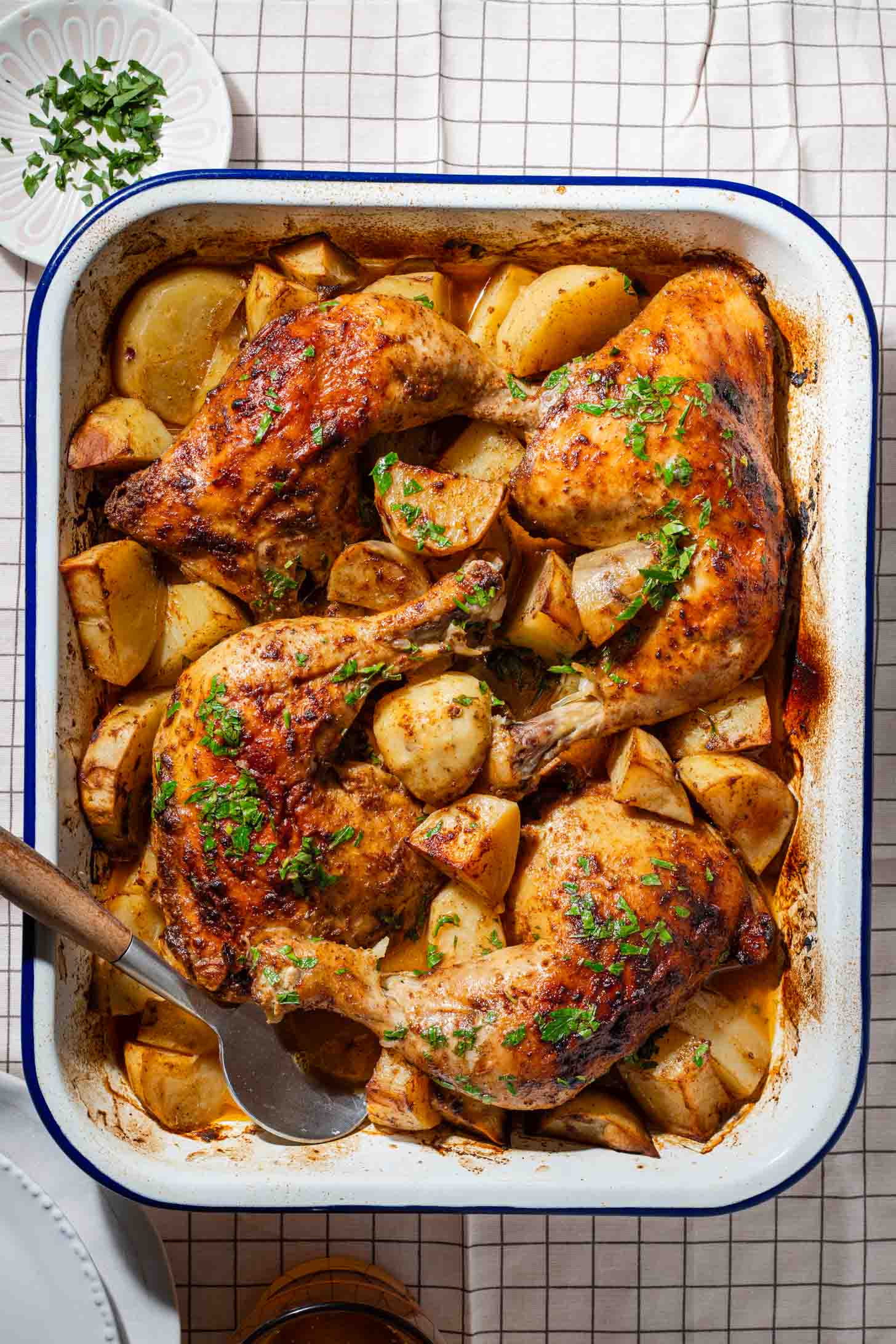 Roasted chicken legs with golden potatoes in a white baking dish, garnished with chopped herbs. A serving spoon and a small plate of extra herbs are nearby on a checkered tablecloth.